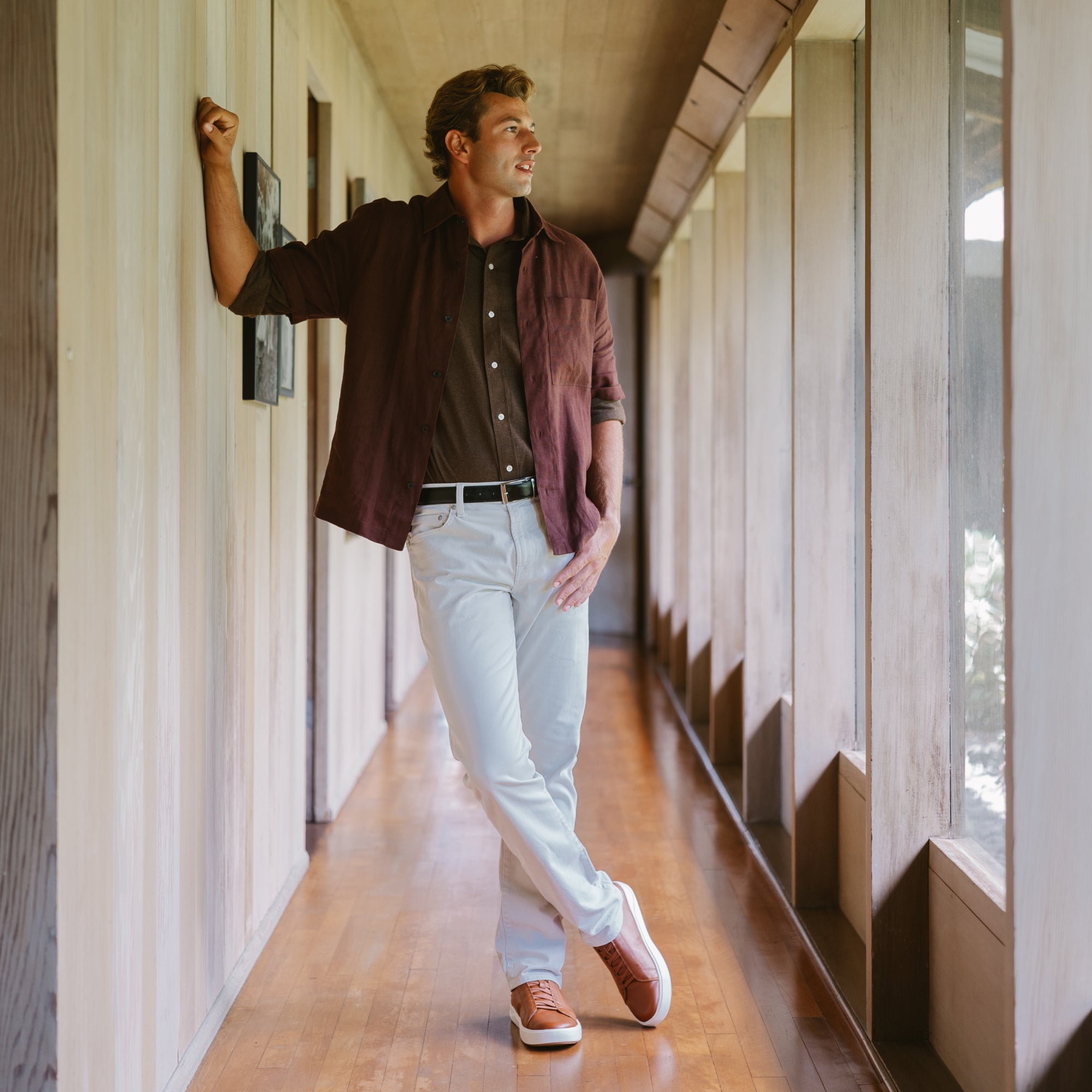 Man wearing dark burgundy button-up shirt over black shirt with light blue pants and brown loafers in a hallway.