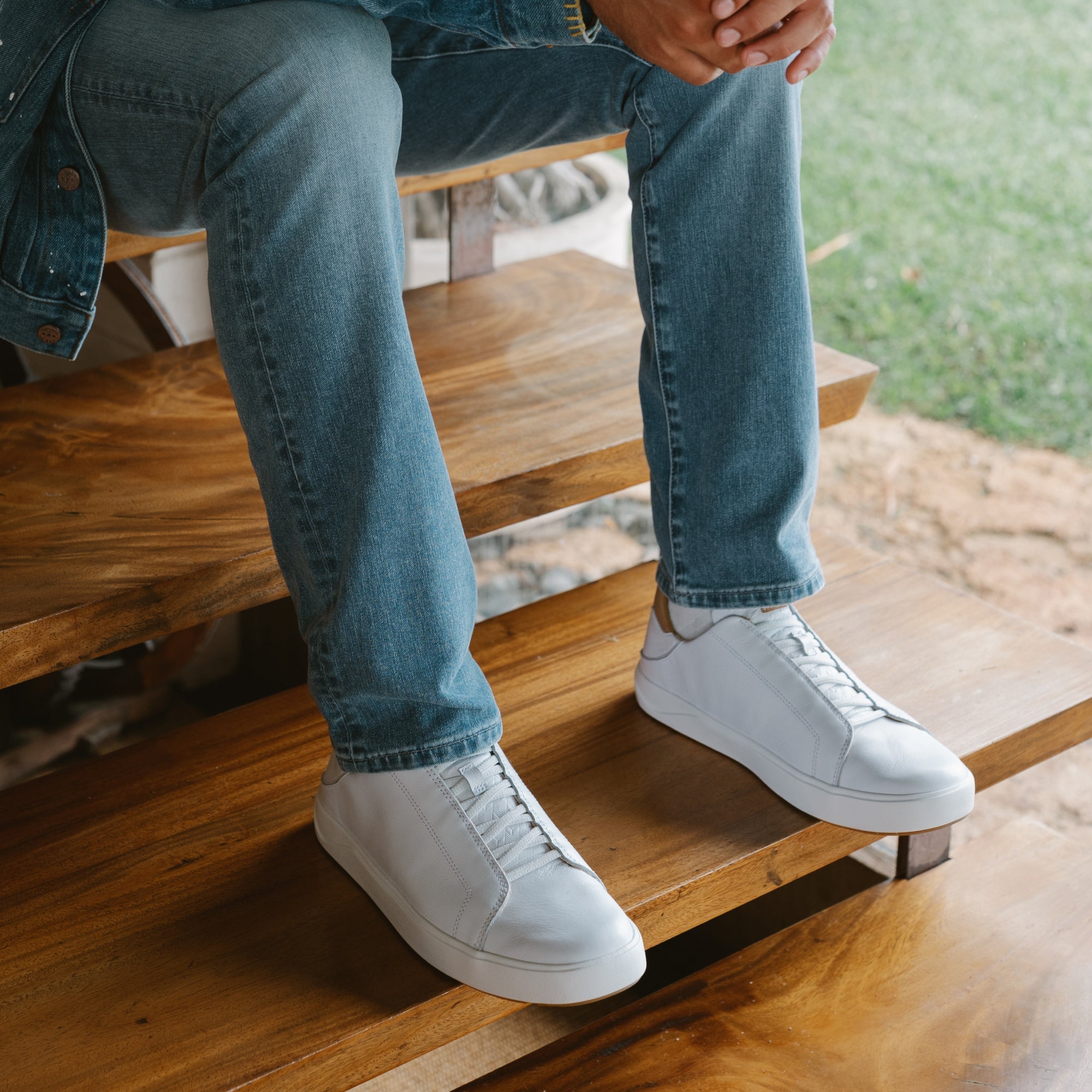 Person wearing bright white low-top sneakers with blue jeans, seated on wooden bench.