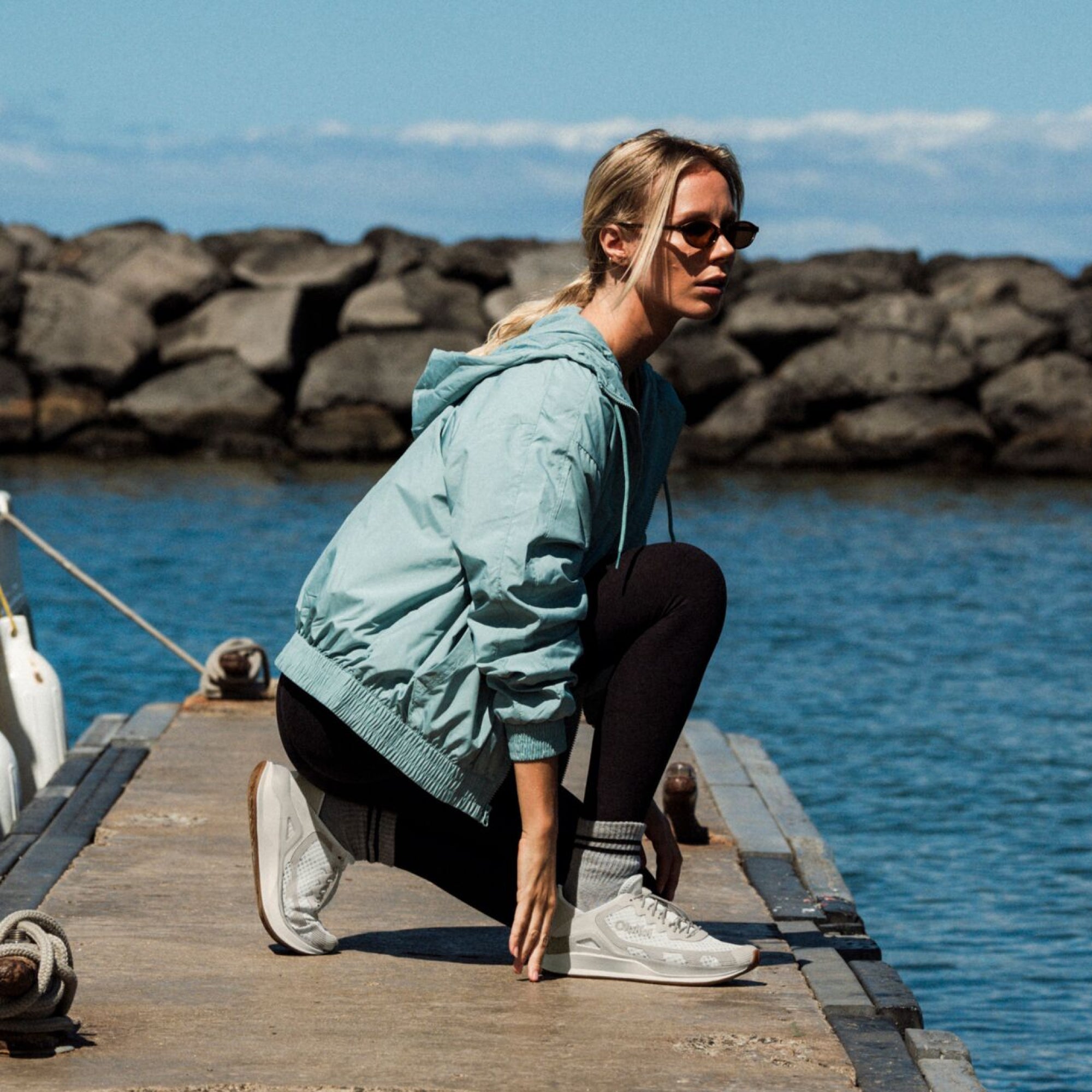 Woman wearing light blue hooded jacket and black pants crouching on wooden dock near water and rocks.