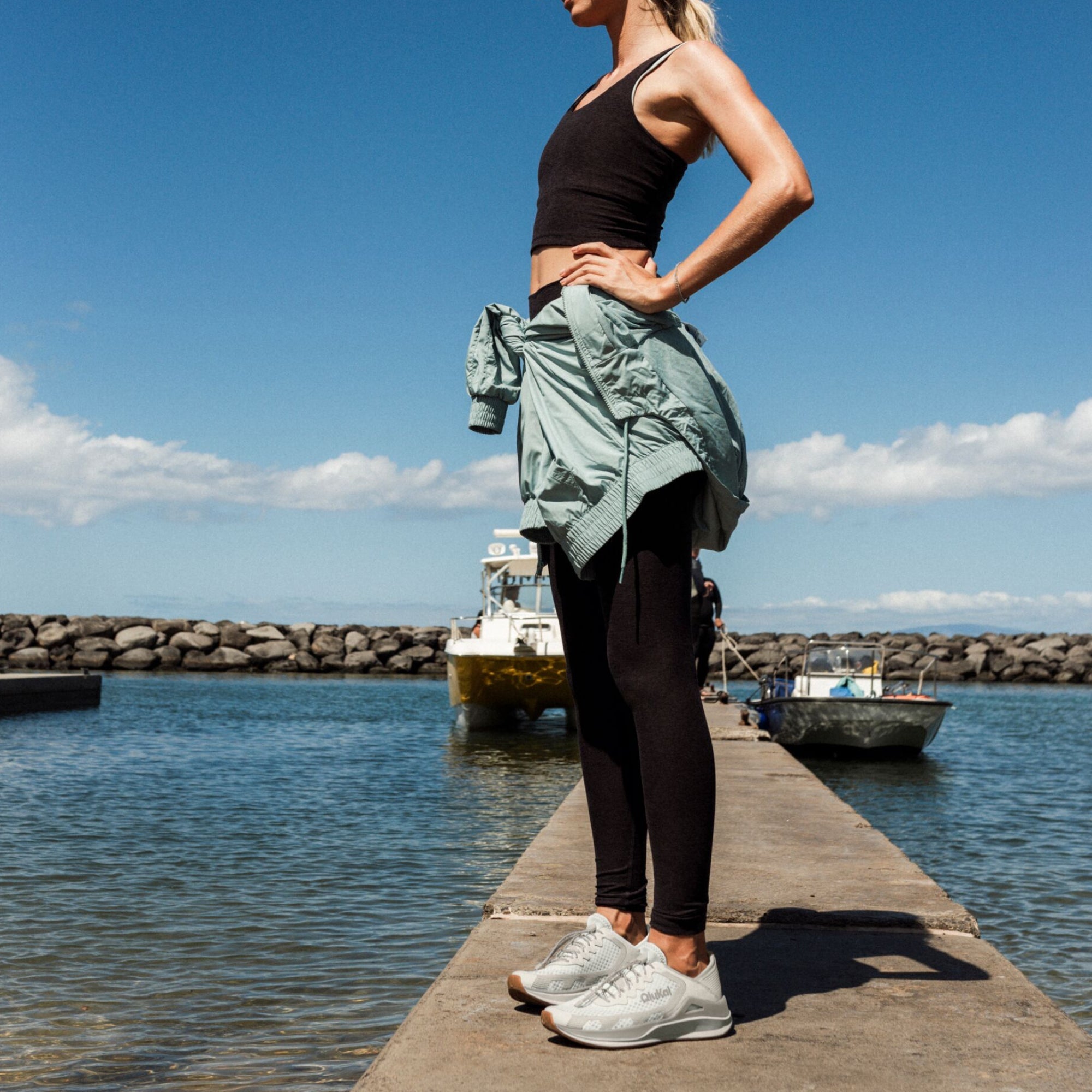 Woman wearing light sage green shirt tied at waist with black tank top and leggings, standing on pier.